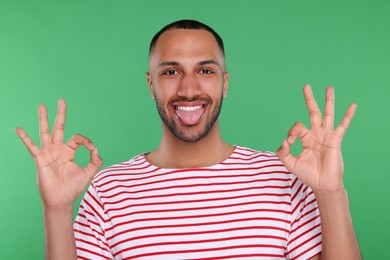 Photo of Happy young man showing his tongue and OK gesture on green background