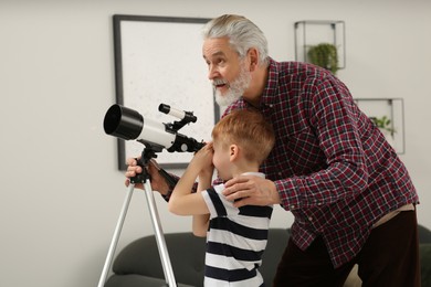 Photo of Little boy with his grandfather looking at stars through telescope in room