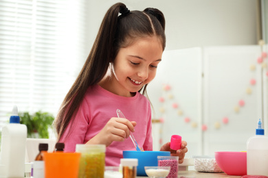 Photo of Cute little girl mixing ingredients with silicone spatula at table. DIY slime toy