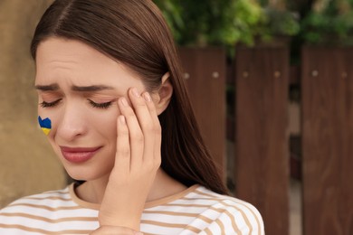 Photo of Sad young woman with drawing of Ukrainian flag on face outdoors, space for text