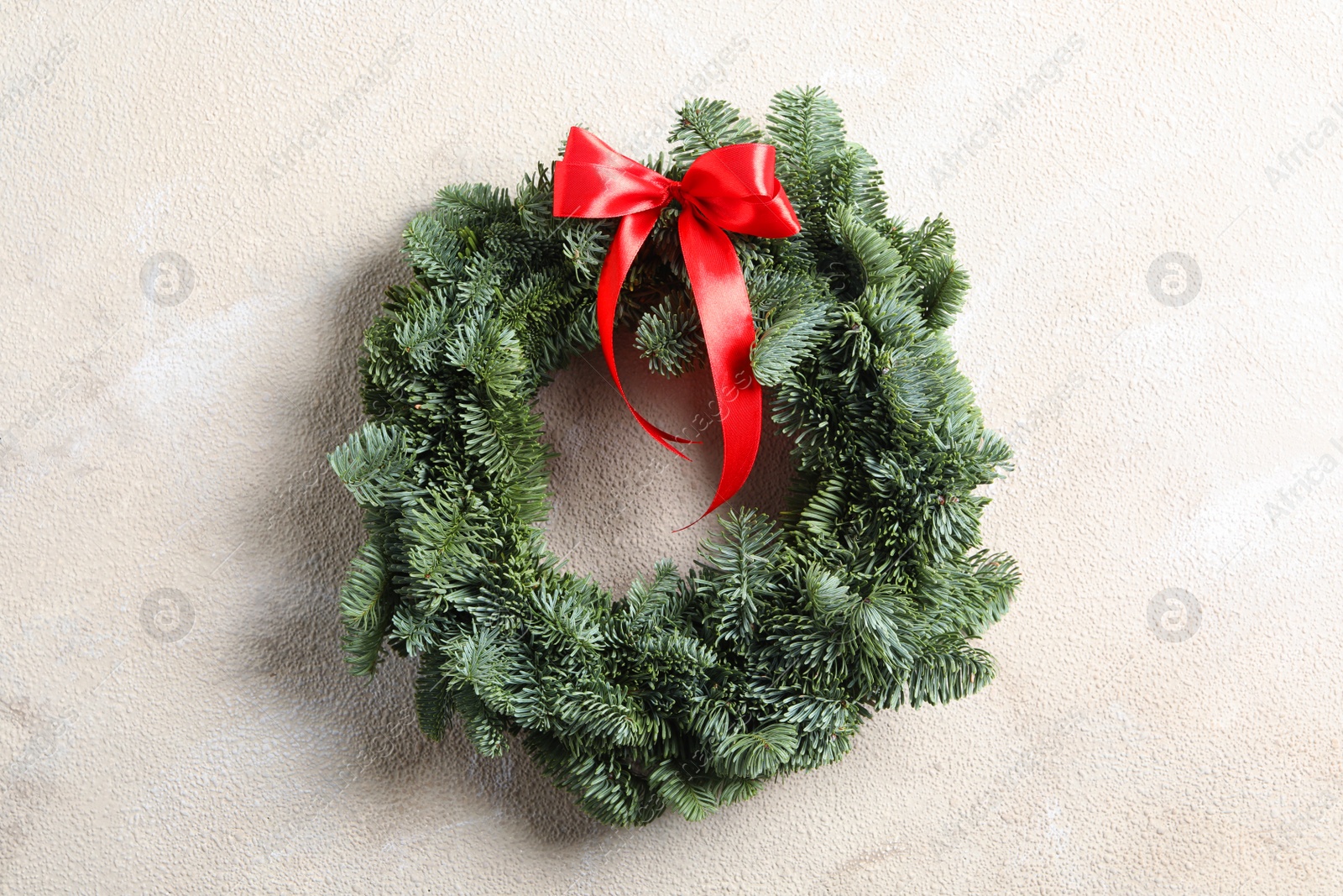 Christmas wreath made of fir tree branches with red ribbon on light textured background Photo of Christmas wreath made of fir tree branches with red ribbon on light textured background
