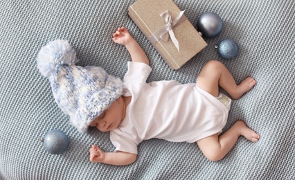 Photo of Cute baby in knitted hat with Christmas decor and gift box sleeping on blanket