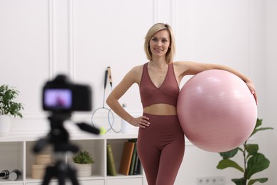 Photo of Smiling sports blogger holding fit ball while recording fitness lesson with camera at home
