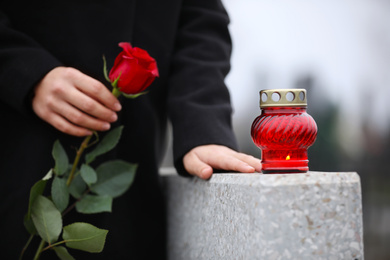 Photo of Woman holding red rose near grey granite tombstone with candle outdoors, closeup. Funeral ceremony