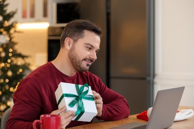 Photo of Celebrating Christmas online with exchanged by mail presents. Man with gift box during video call on laptop at home