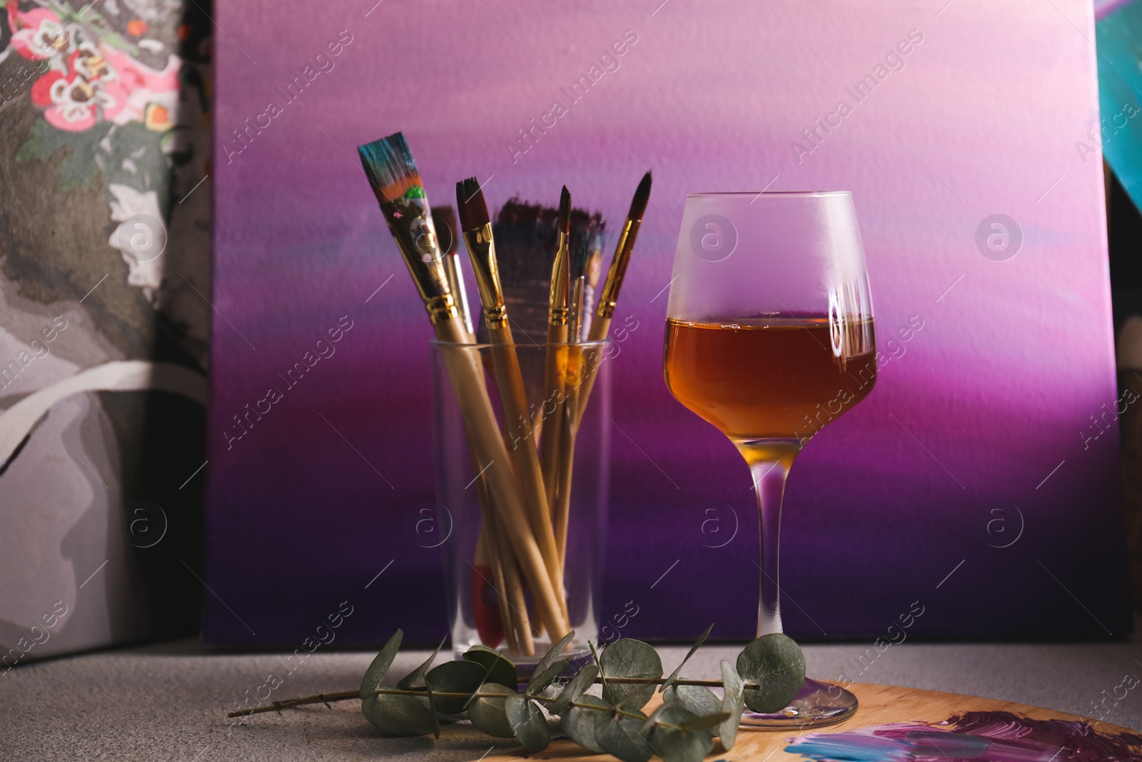 Photo of Glass of tasty wine, brushes with colorful paints and gradient canvas on light gray table