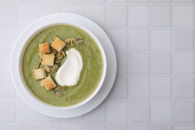 Photo of Delicious broccoli cream soup with croutons, sour cream and pumpkin seeds on white tiled table, top view. Space for text