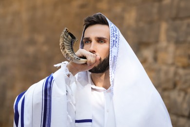 Photo of Jewish man blowing shofar on Rosh Hashanah outdoors. Wearing tallit with words Blessed Are You, Lord Our God, King Of The Universe, Who Has Sanctified Us With His Commandments, And Commanded Us To Enwrap Ourselves In Tzitziton in Hebrew