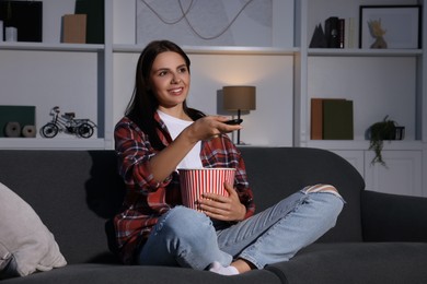 Photo of Happy woman holding popcorn bucket and changing TV channels with remote control at home