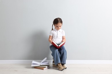 Photo of Cute little girl reading on stack of books near light grey wall