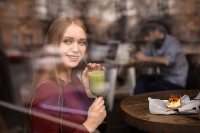 Pretty young woman with cocktail and cake at table in cafe, view from outdoors through window