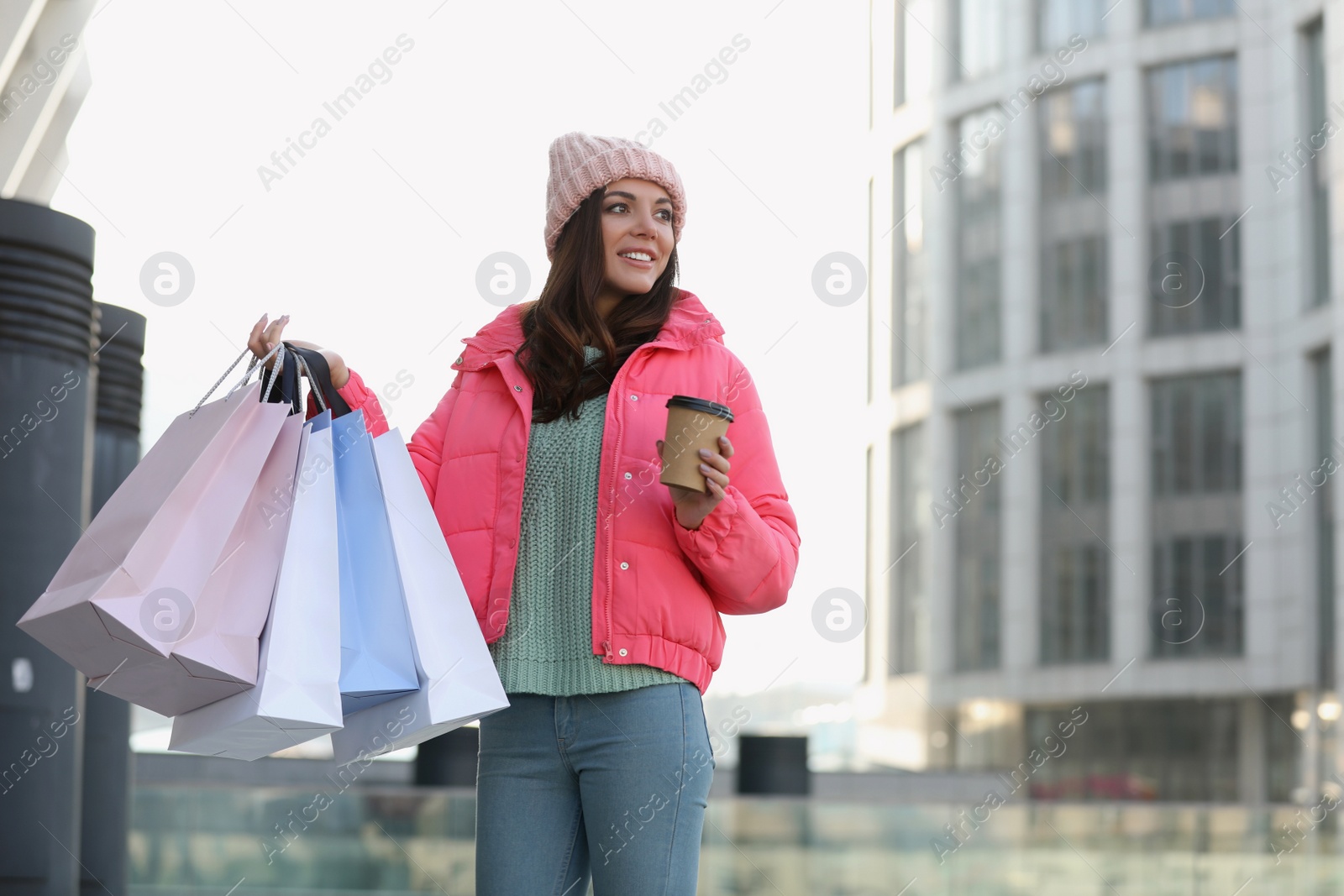 Photo of Beautiful young woman with cup of coffee and shopping bags near building