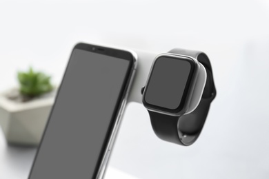 Photo of Stylish smart watch and phone charging with wireless pad against blurred background, closeup