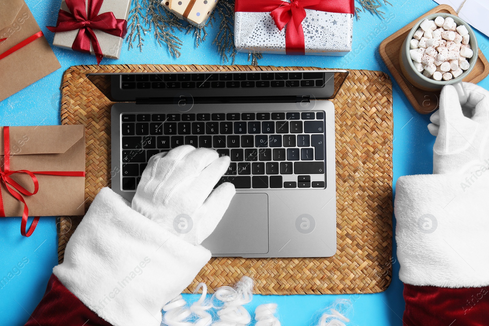 Photo of Santa Claus using laptop, closeup. Gift boxes, cup of hot drink and Christmas decor on light blue background, top view