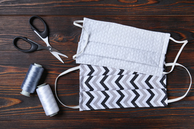 Homemade protective masks and sewing accessories on wooden background, flat lay