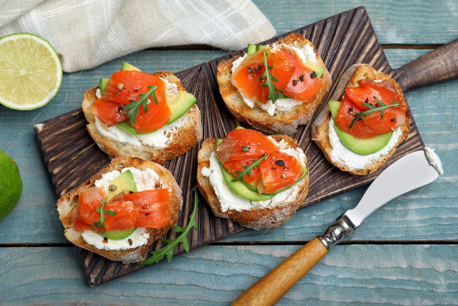 Photo of Delicious sandwiches with cream cheese, salmon, avocado and arugula served on light blue wooden table, flat lay