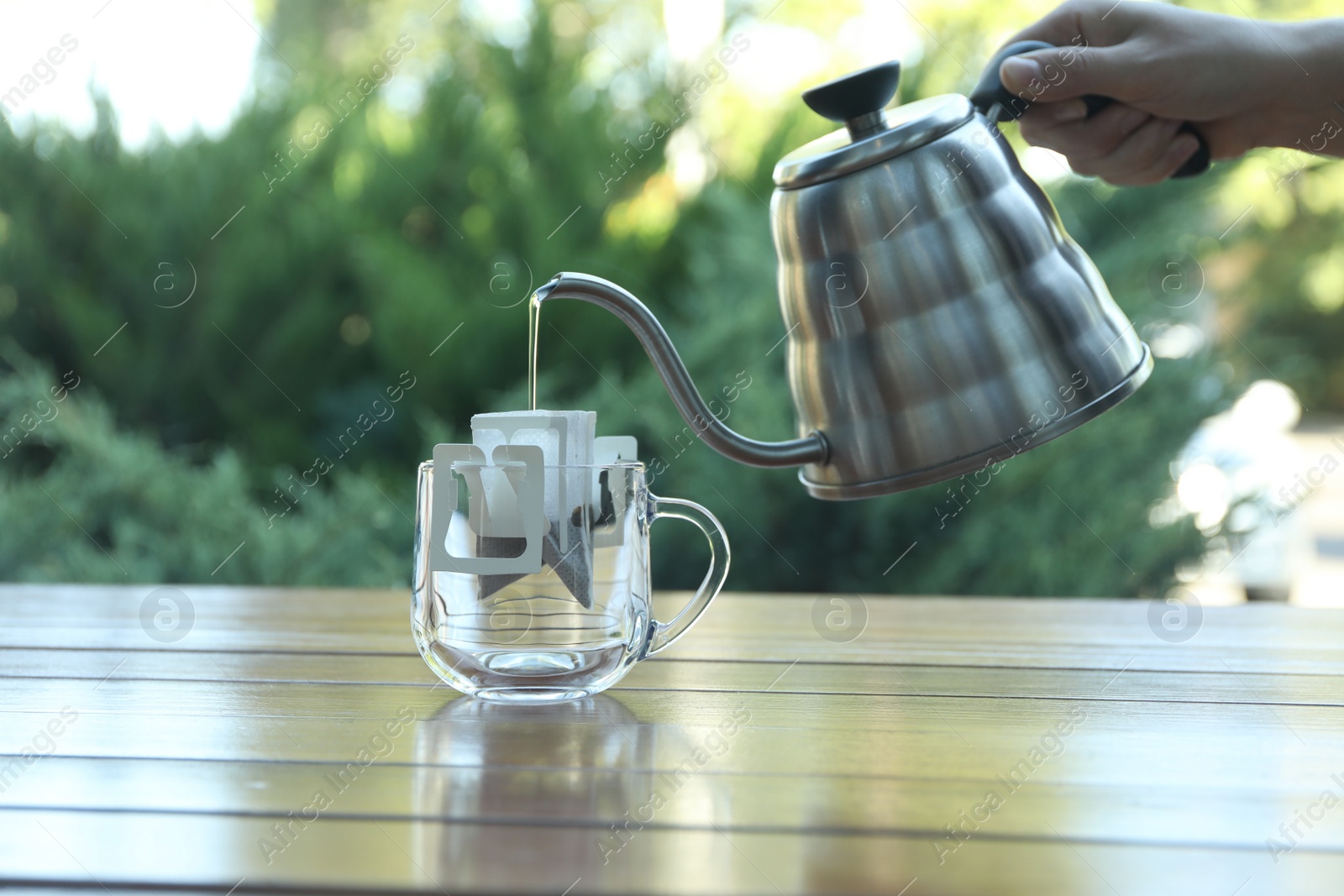 Photo of Woman pouring hot water into glass cup with drip coffee bag from kettle at wooden table, closeup