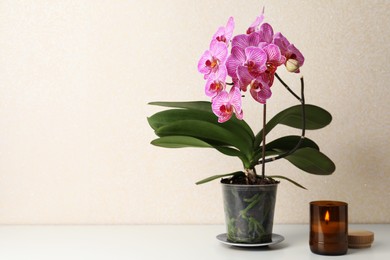 Photo of Beautiful blooming orchid and burning candle on white table near beige wall. Space for text