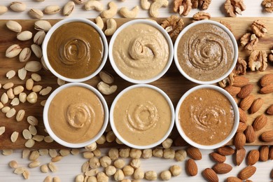 Many tasty nut butters in bowls and nuts on white table, flat lay