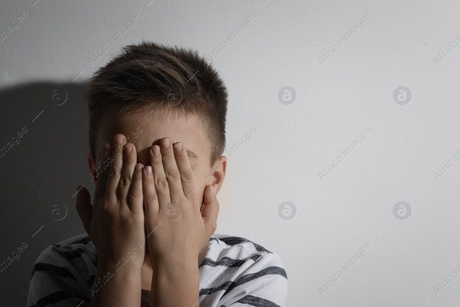 Photo of Scared little boy closing his eyes near white wall, space for text. Domestic violence concept