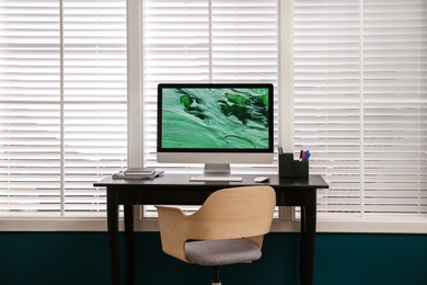 Photo of Comfortable workplace near window with white horizontal blinds in room