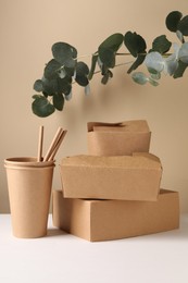 Photo of Eco friendly food packaging. Paper containers, straws and eucalyptus branches on white table against beige background