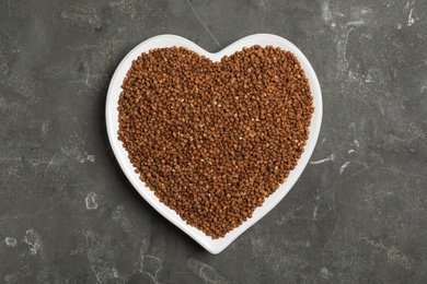 Heart shaped plate with raw buckwheat on grey background, top view