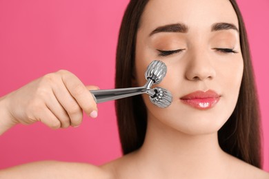 Woman using metal face roller on pink background, closeup