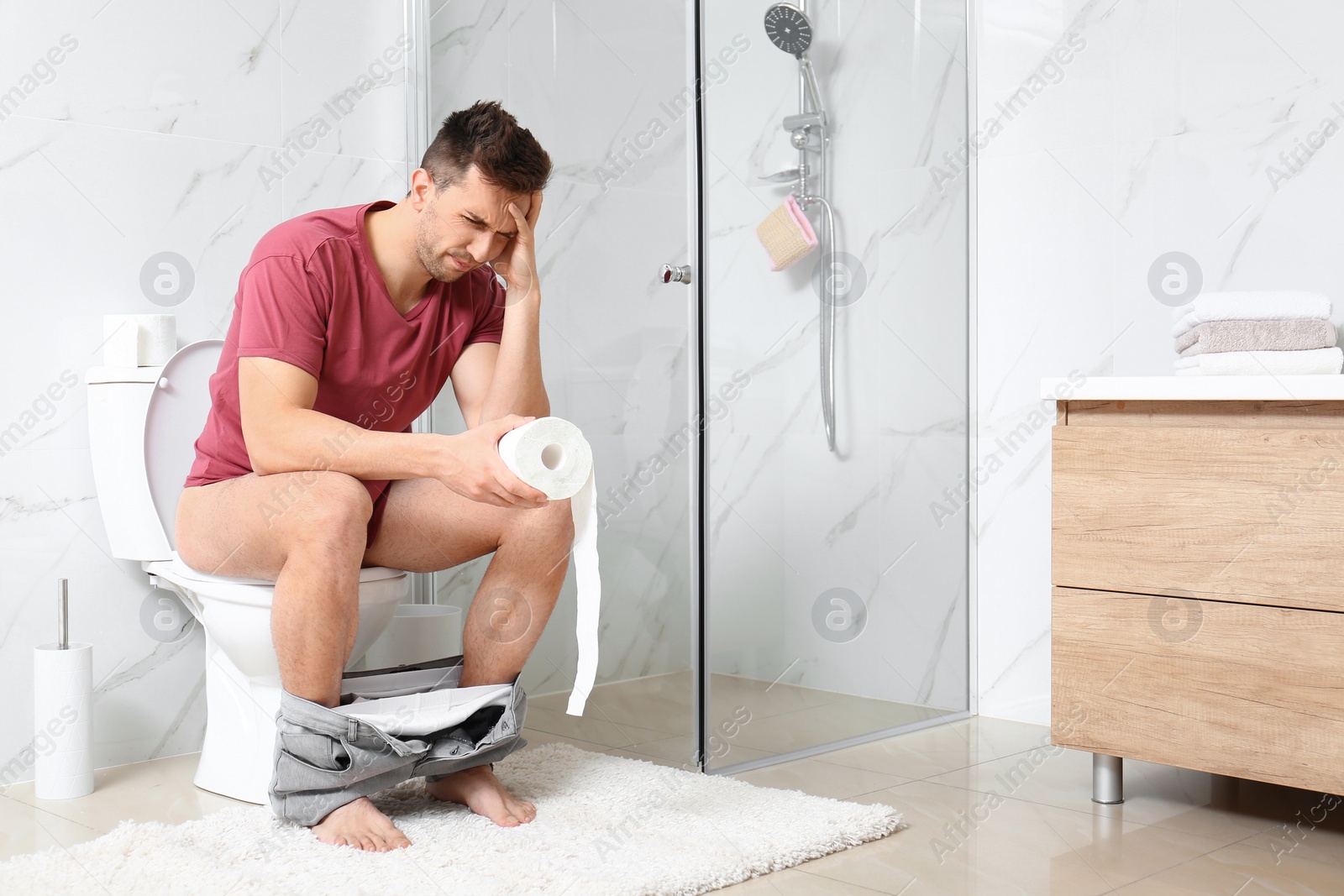 Photo of Man with paper roll suffering from stomach ache on toilet bowl in bathroom