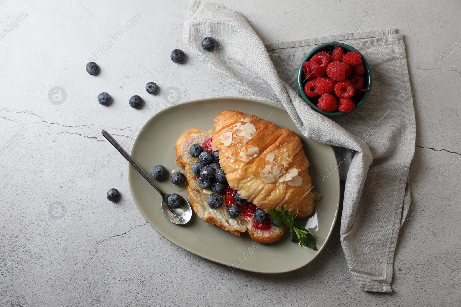 Photo of Delicious croissant with berries, almond flakes and spoon on grey table, flat lay