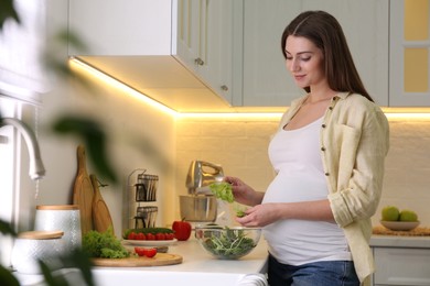 Young pregnant woman preparing vegetable salad at table in kitchen. Healthy eating