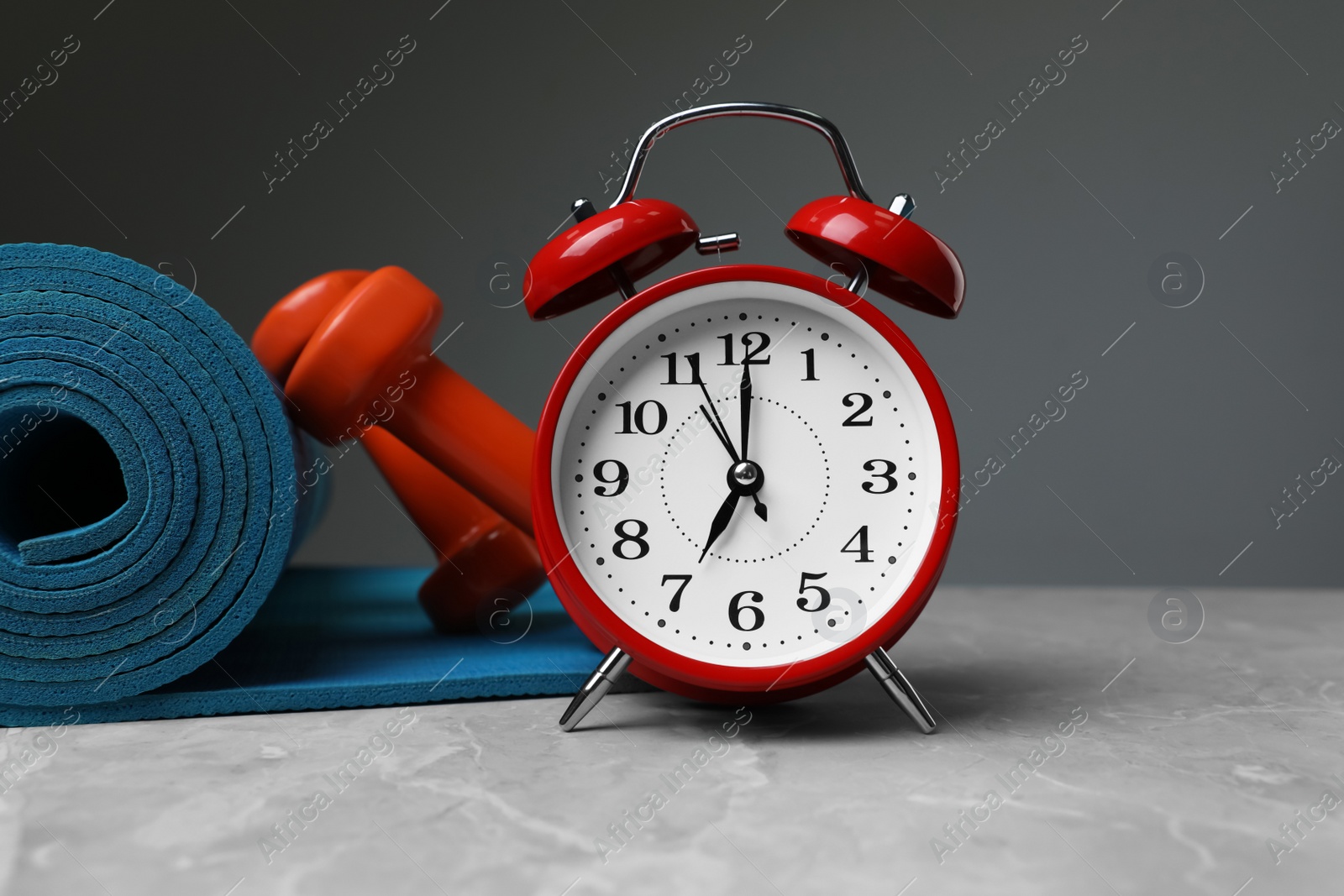 Photo of Alarm clock, dumbbells and yoga mat on table against grey background. Morning exercise