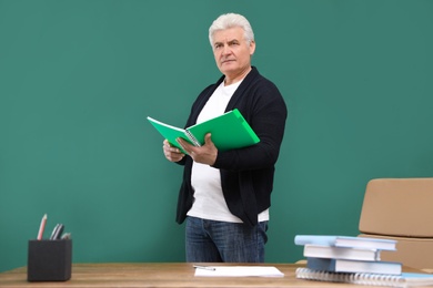 Portrait of senior teacher with notebook at green chalkboard and table