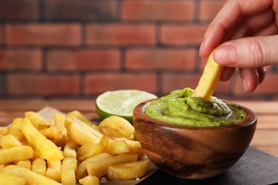 Photo of Woman dipping french fry into avocado sauce at wooden table, closeup