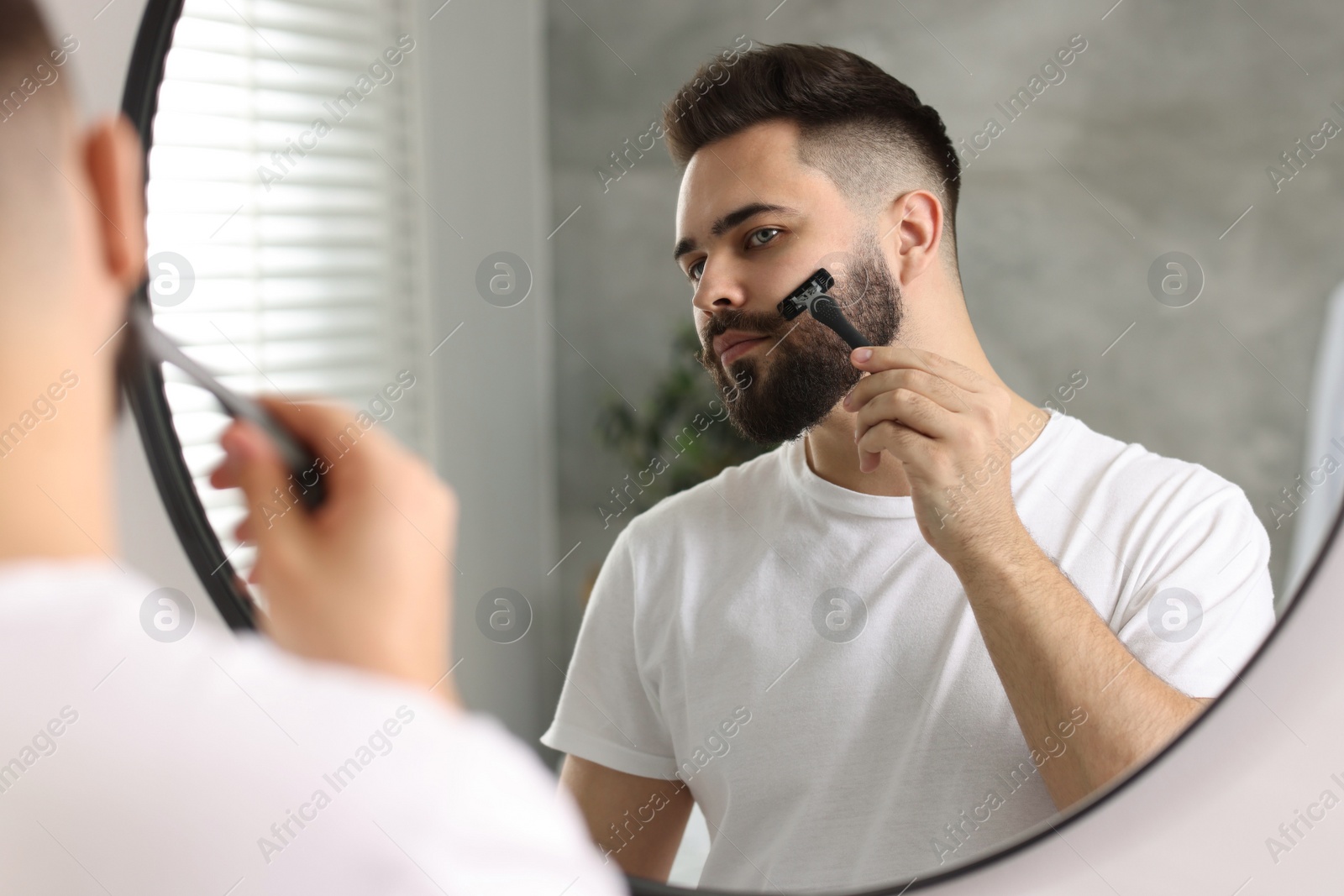 Photo of Handsome young man shaving with razor near mirror in bathroom