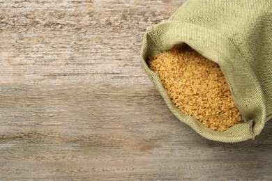 Burlap bag with uncooked bulgur on wooden table, top view. Space for text