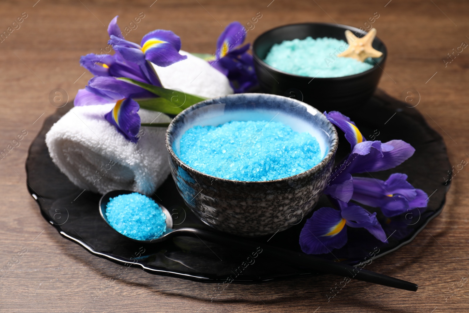 Photo of Light blue sea salt in bowls, flowers, towel and spoon on wooden table