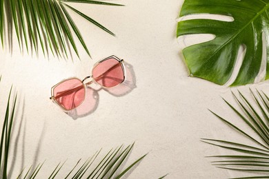 Stylish sunglasses and different tropical leaves on sand, flat lay