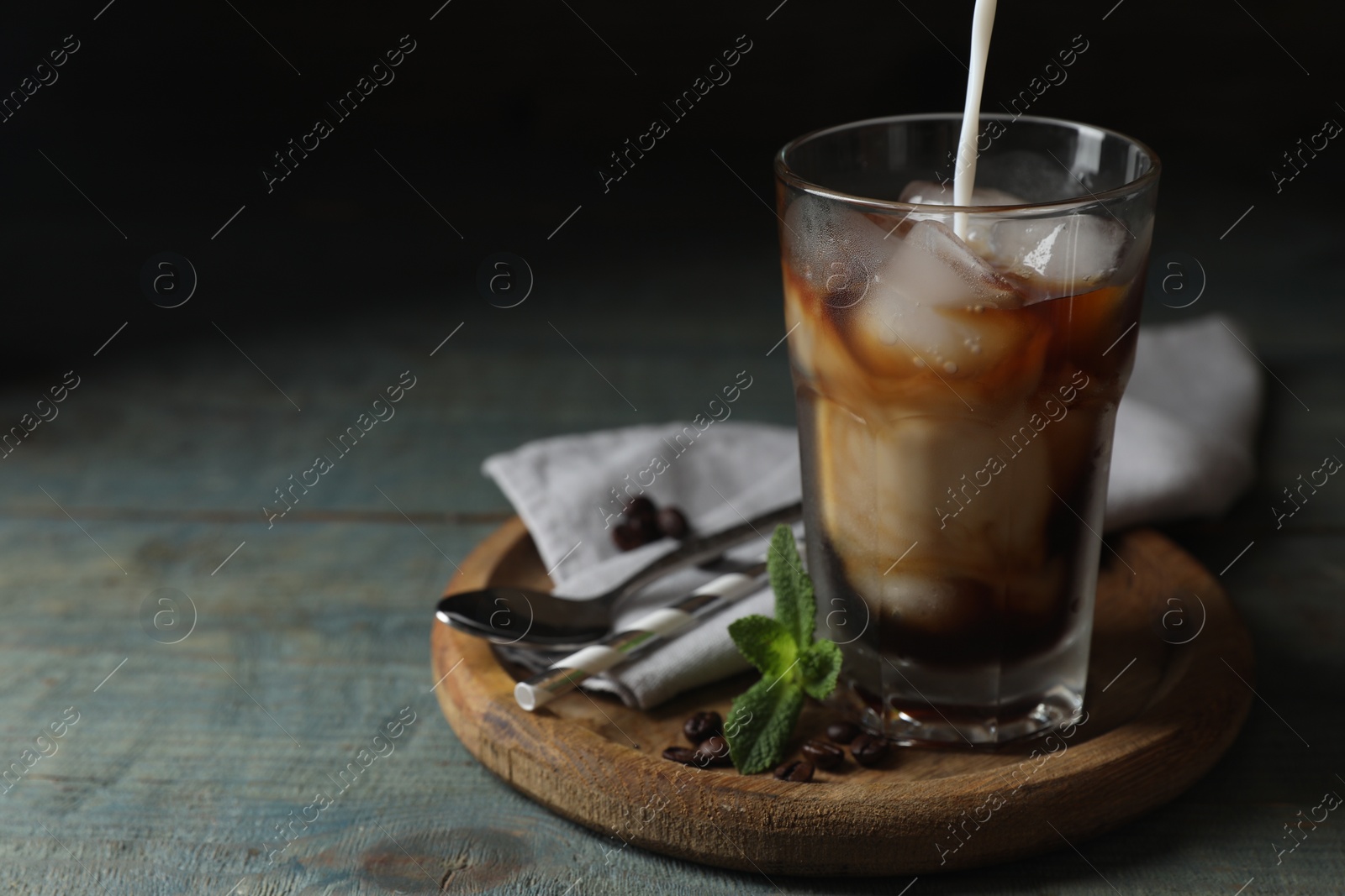 Photo of Pouring milk into glass of delicious iced coffee, mint and beans on light blue wooden table, space for text