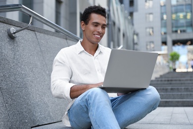 Handsome young African-American man with laptop sitting on stairs outdoors