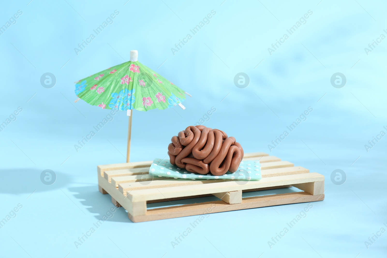 Photo of Brain made of plasticine on mini wooden sunbed under umbrella against light blue background