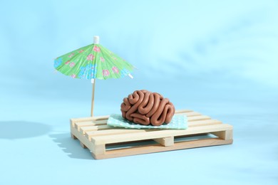 Photo of Brain made of plasticine on mini wooden sunbed under umbrella against light blue background