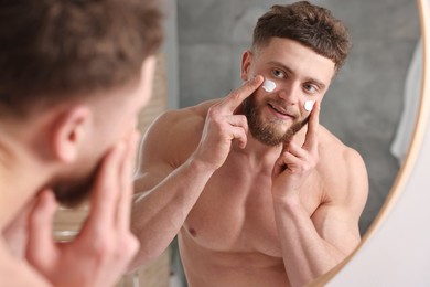 Handsome man applying moisturizing cream onto his face near mirror in bathroom
