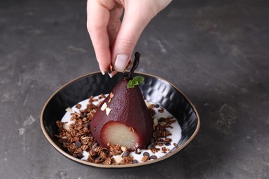 Woman putting muesli on tasty red wine poached pear in bowl at grey table, closeup