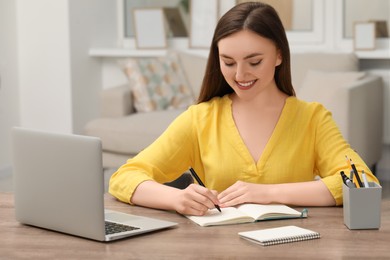 Photo of Young woman writing in notebook while working on laptop at wooden table indoors