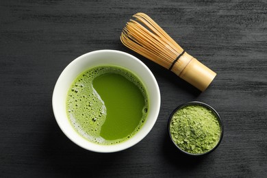 Photo of Cup of fresh matcha tea, green powder and bamboo whisk on black wooden table, flat lay