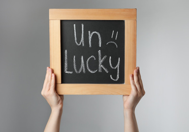 Photo of Woman holding blackboard with word UNLUCKY on light grey  background, closeup