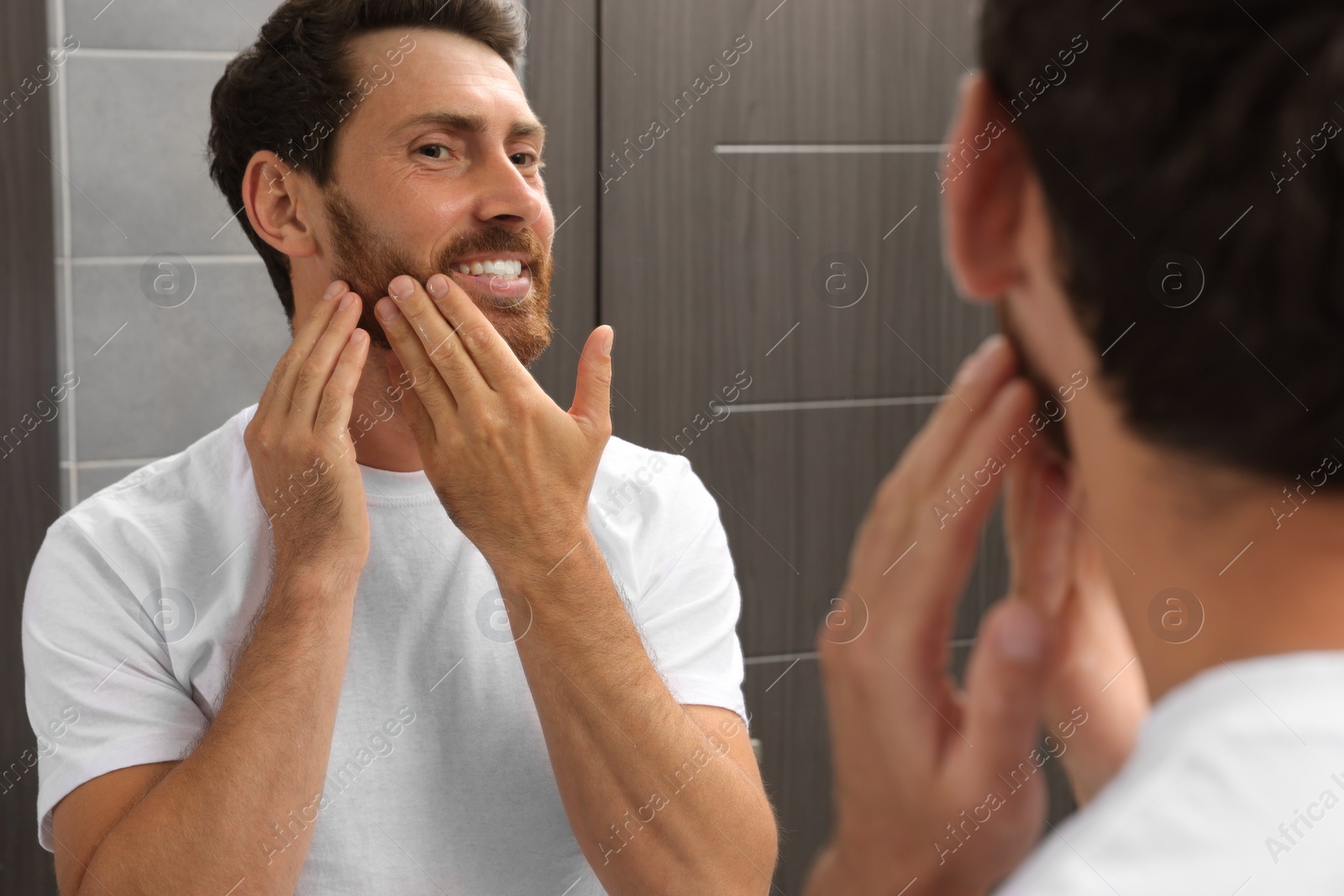 Photo of Smiling bearded man looking at mirror in bathroom