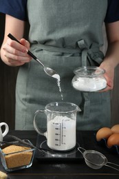 Photo of Woman adding baking powder into measuring cup at black wooden table, closeup