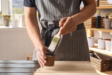 Photo of Man dipping brush into can of wood stain at wooden surface indoors, closeup